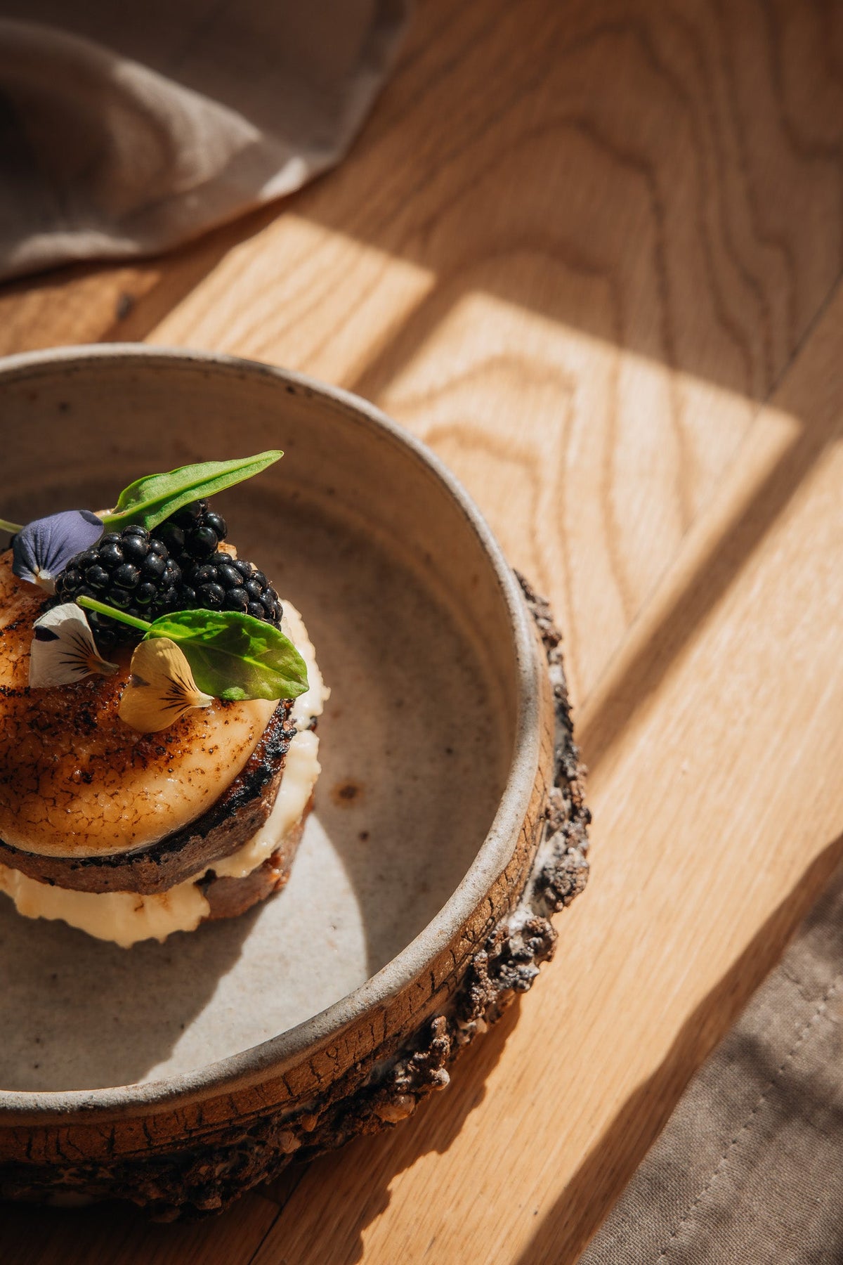 A gourmet dessert or pastry topped with blackberries, green leaves, and edible flowers, served on a rustic ceramic plate with a bark edge, set on a wooden table in warm natural light.