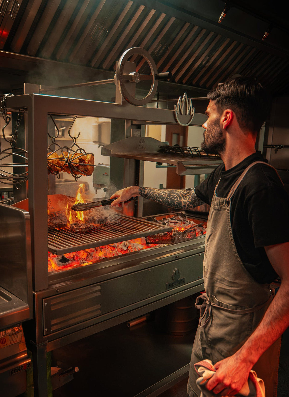 A professional chef in a dark kitchen, wearing a gray apron and black t-shirt, actively cooking meat on a large stainless steel rotisserie grill with visible flames and smoke.