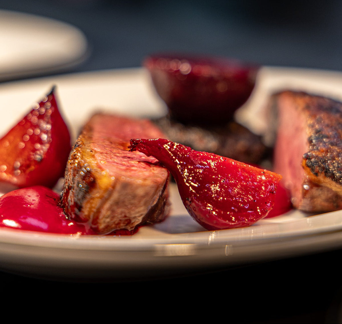 A close-up shot of a plated gourmet dish featuring sliced medium-rare steak with charred edges, served with glossy red pear slices and a dark berry sauce on a white plate against a dark, blurred background.