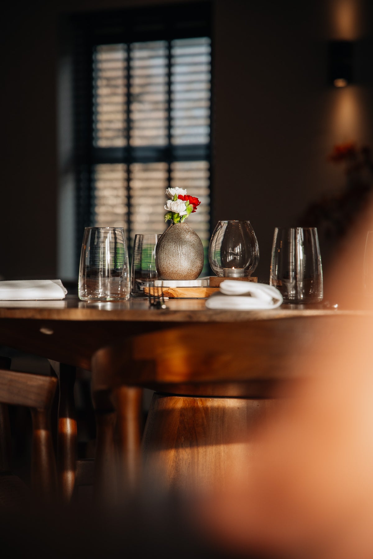 A warmly lit dining table setting with clear wine glasses, white napkins, a small round vase with red and white flowers, and wooden chairs in a softly blurred indoor environment.